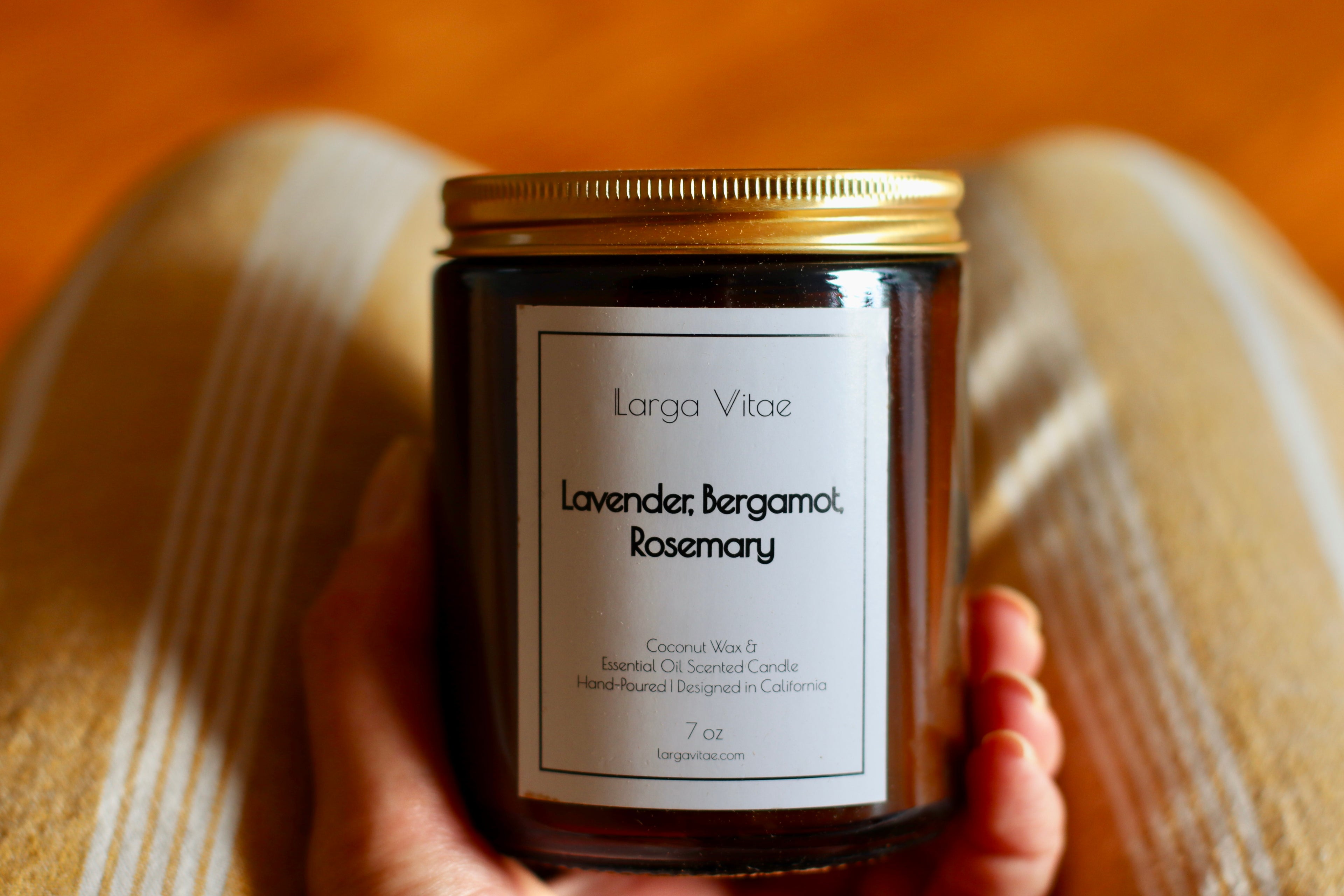 Lavender, Bergamot, Rosemary essential oil scented candle jar by Larga Vitae in a woman's hand on her pinstriped yellow panted lap