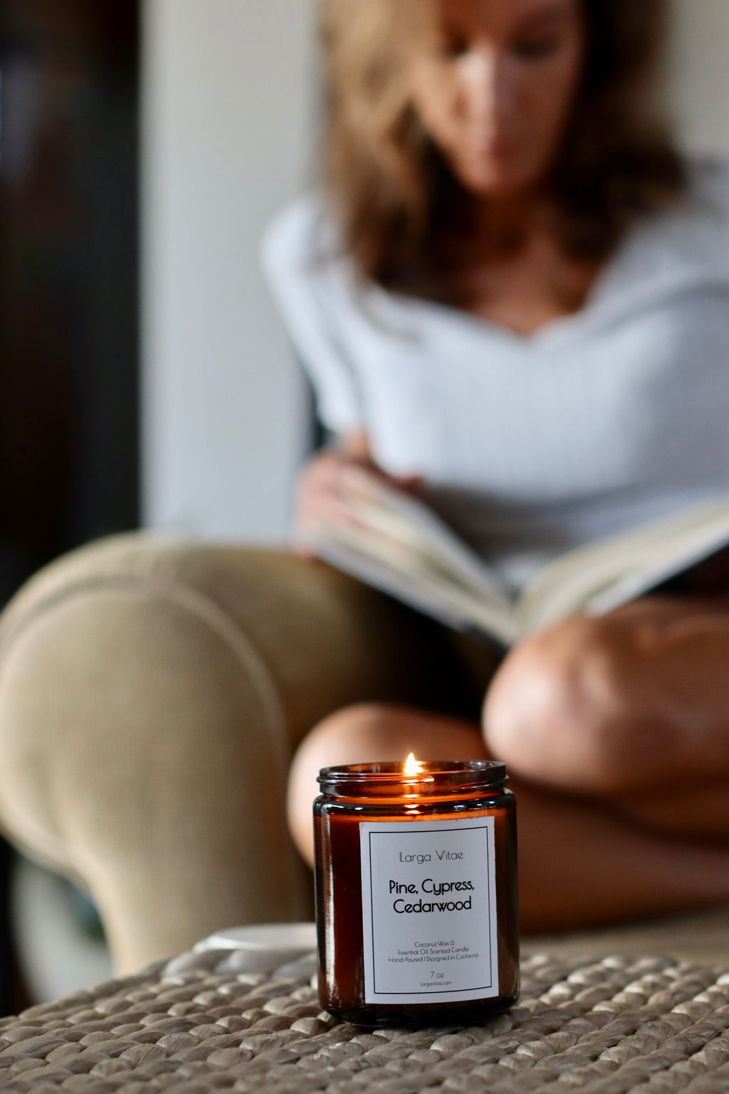 Person reading a book with a lit Larga Vitae pine, cypress, cedarwood candle on a table in the foreground