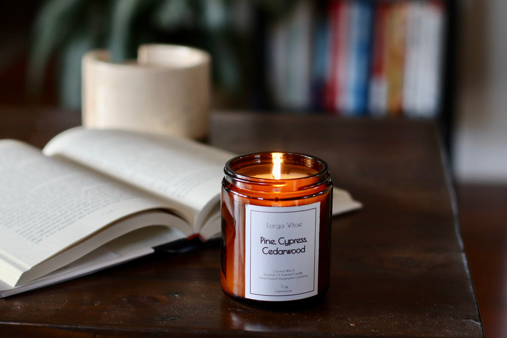 Larga Vitae's Pine, Cypress, Cedarwood coconut wax candle burning on a wooden surface next to an open book