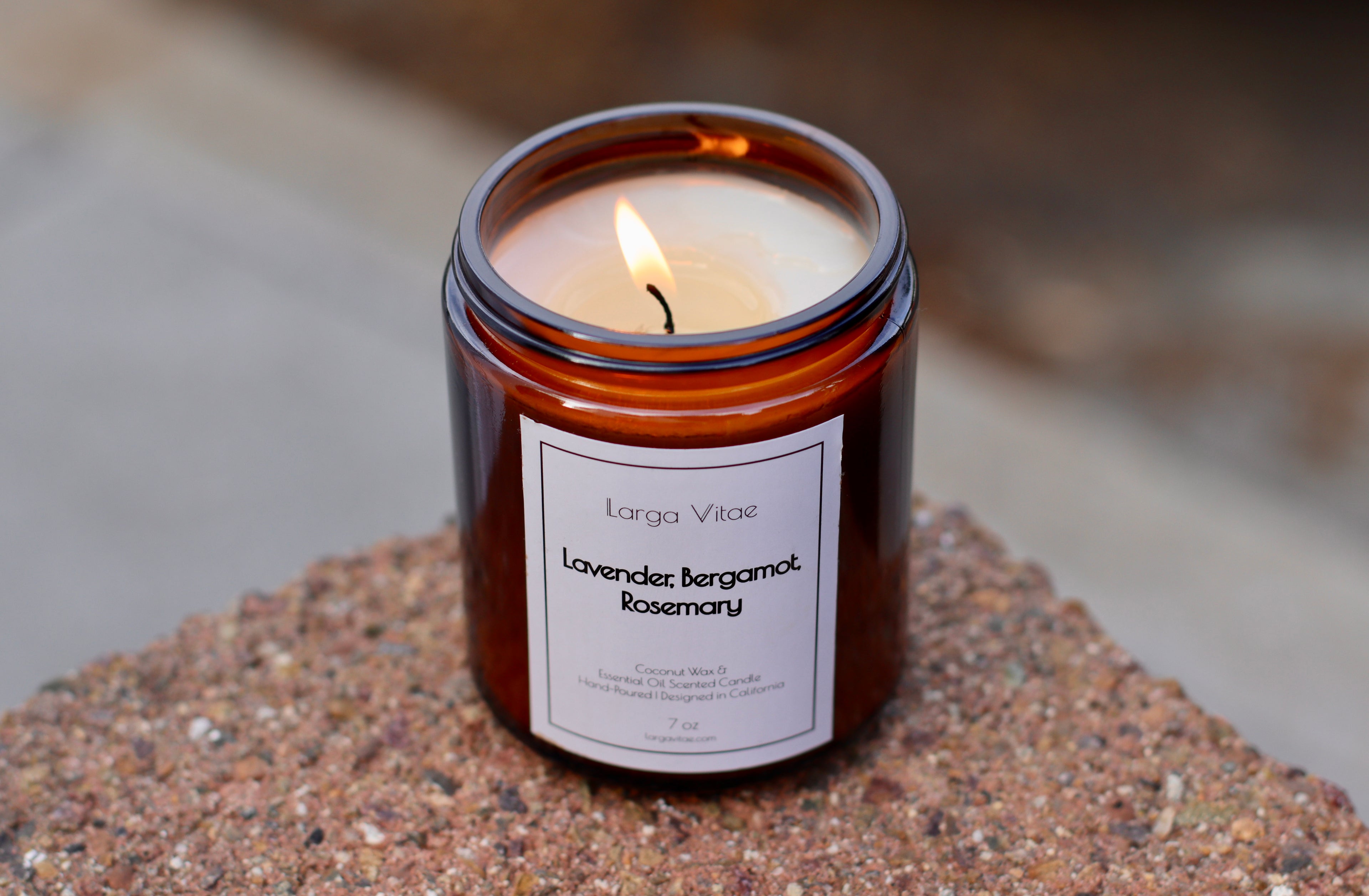 Lit Lavender, Bergamot, Rosemary Larga Vitae natural coconut wax candle in a brown jar on a textured surface