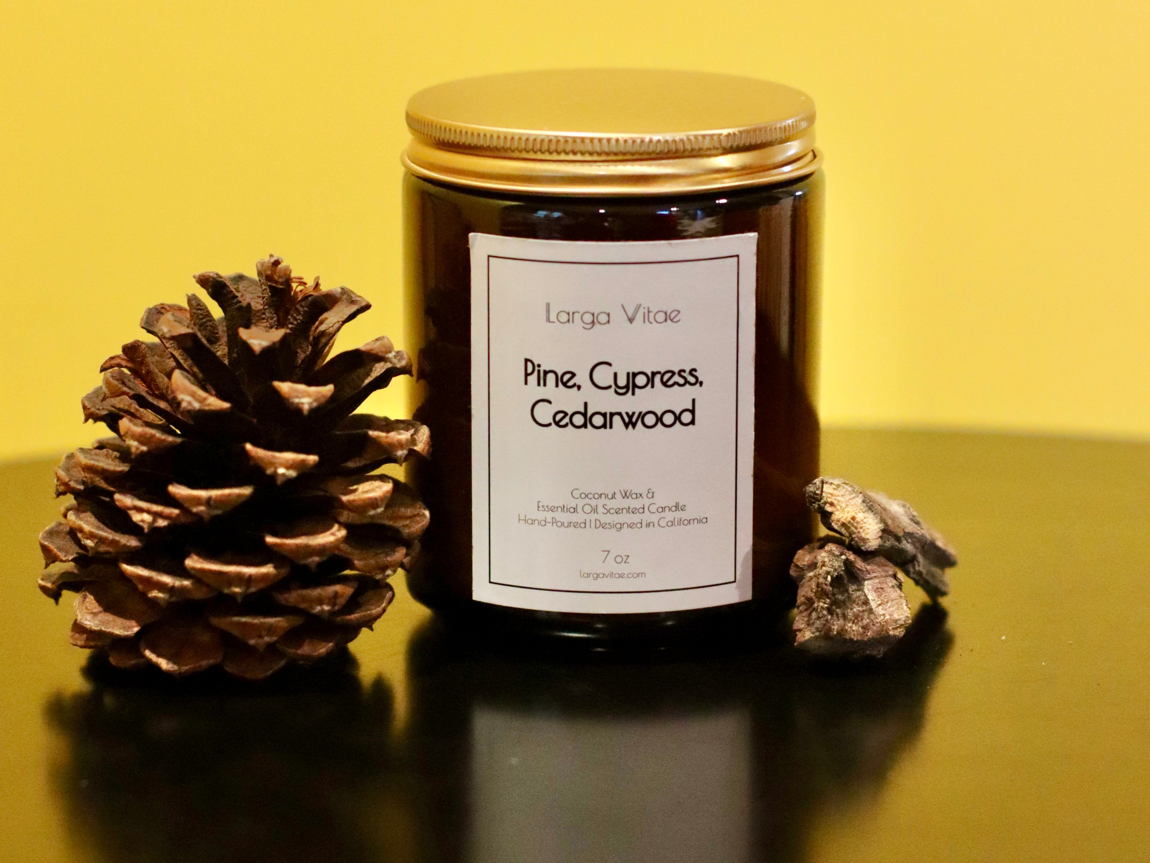 Larga Vitae's Pine, Cypress, Cedarwood jar candle with a pine cone and cedarwood chip on a yellow background