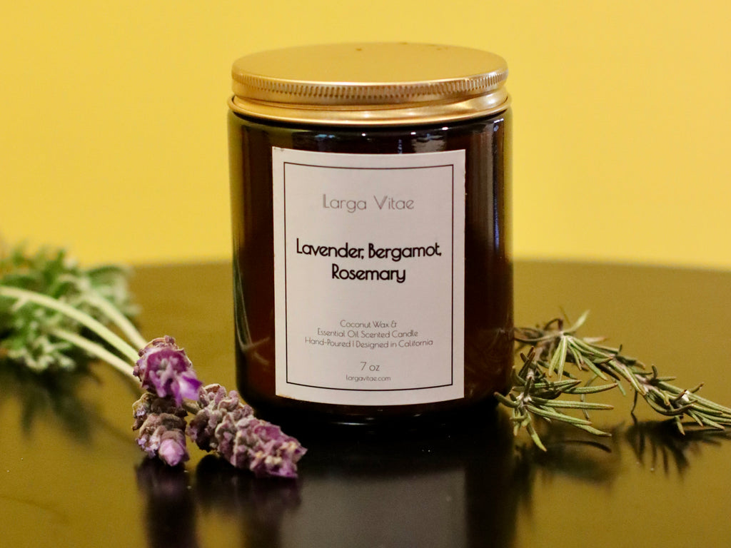7oz candle jar labeled 'Lavender Bergamot Rosemary' by Larga Vitae on a reflective surface with lavender and rosemary sprigs in front of a yellow background.