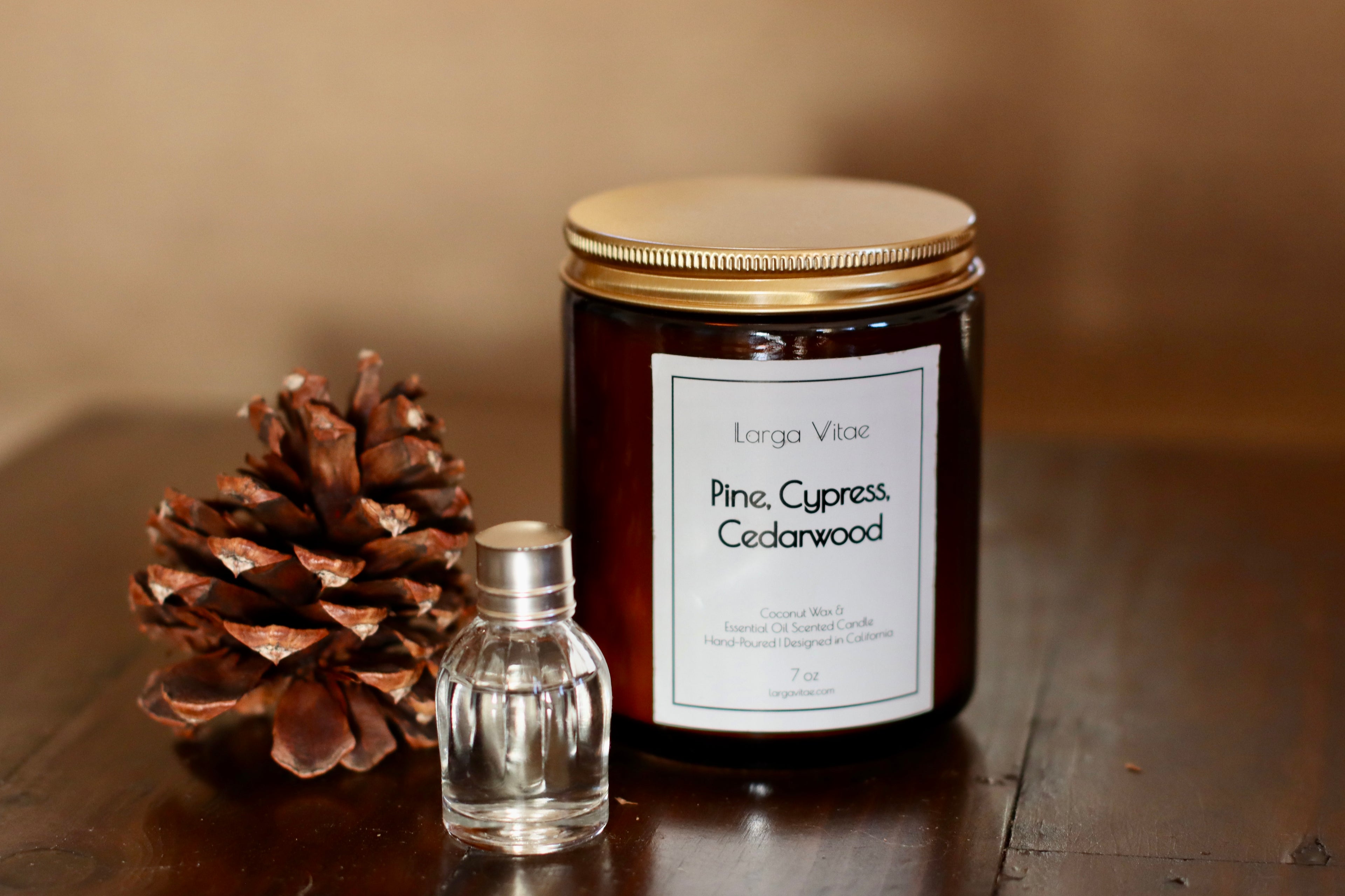 Larga Vitae's Pine, Cypress, Cedarwood natural fragrance oil coconut wax candle with a pine cone and small decorative bottle of essential oil on a wooden surface.