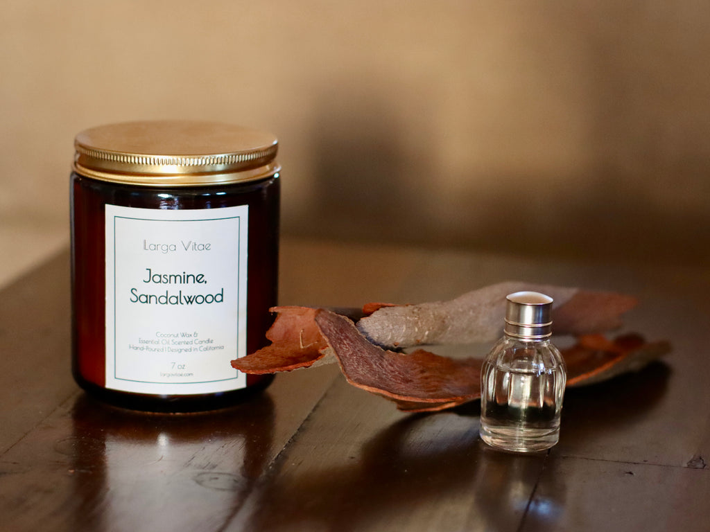 Larga Vitae's Jasmine, Sandalwood Candle with a small bottle of essential oil, and sandalwood pieces on a wooden surface.
