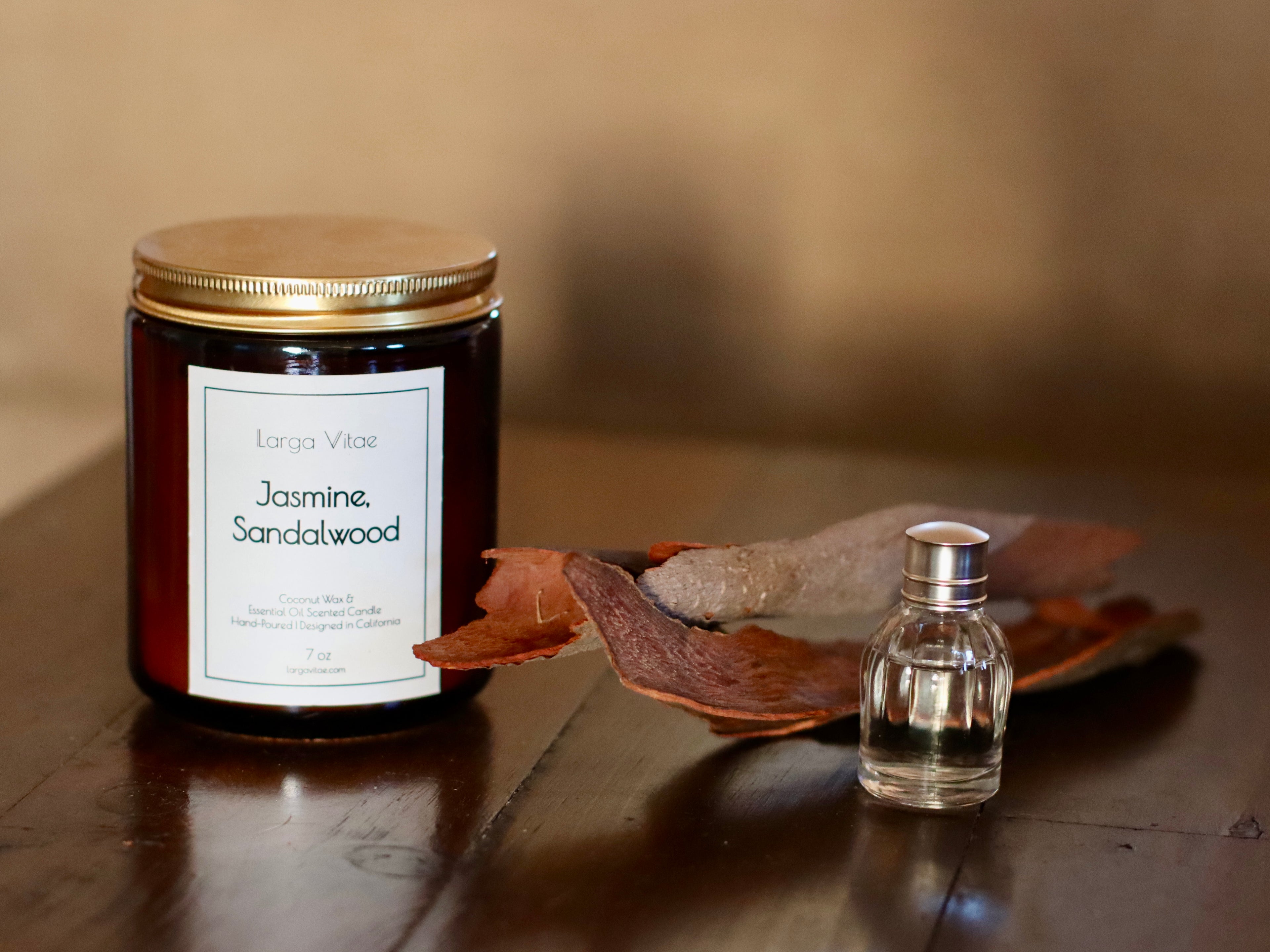 Larga Vitae's Jasmine, Sandalwood Candle with a small bottle of essential oil, and sandalwood pieces on a wooden surface.