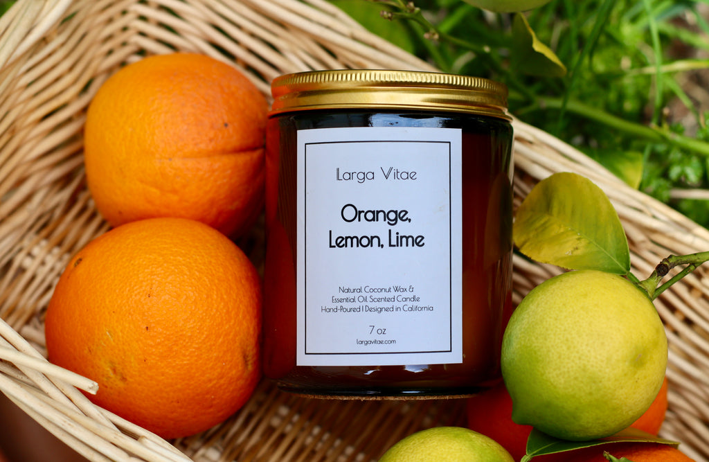 Natural coconut wax candle labeled 'Orange, Lemon, Lime' by Larga Vitae in a basket with oranges and a lemon.