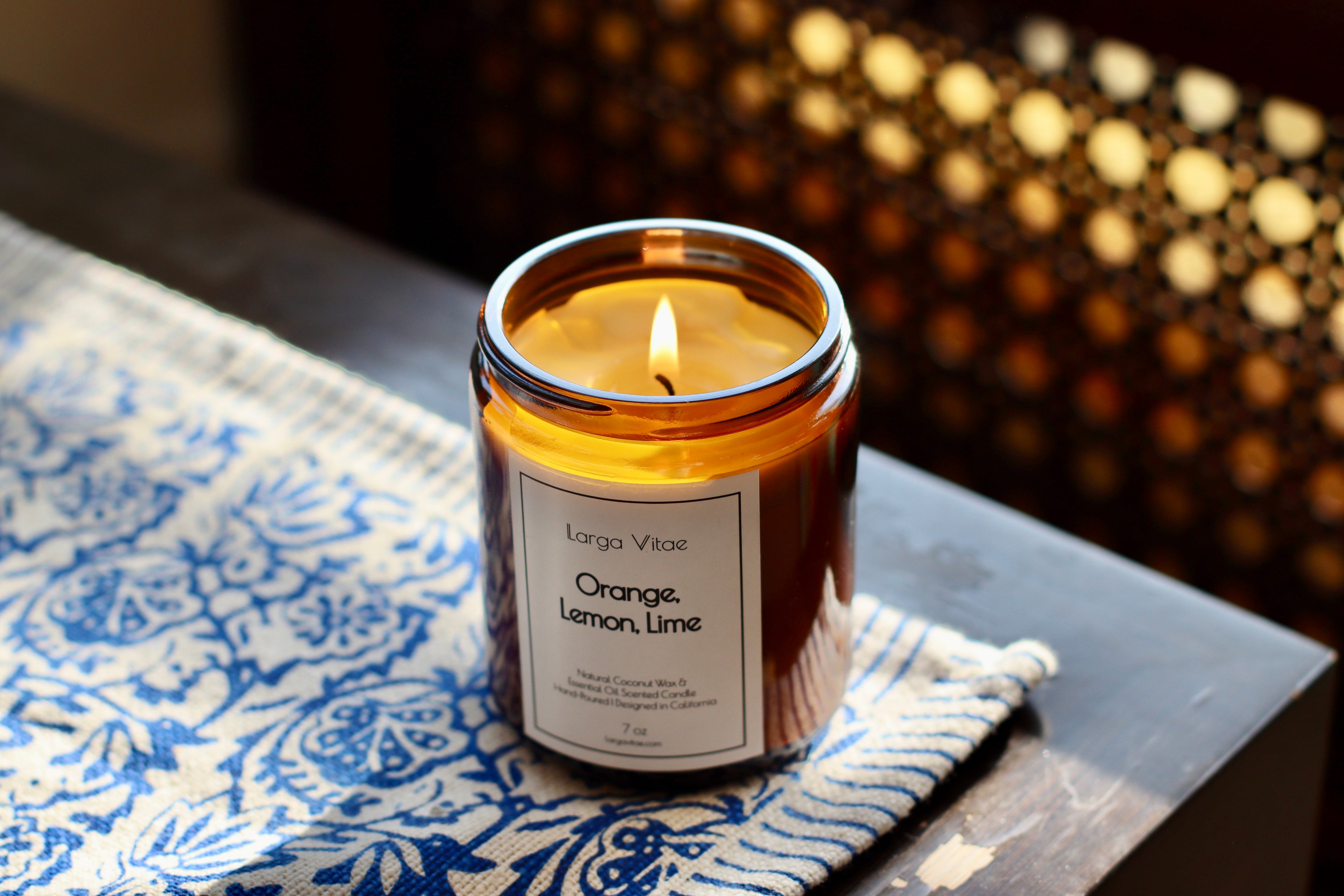 Lit refreshing essential oil candle coconut wax labeled 'Orange Lemon Lime' on a patterned fabric with a blurred background