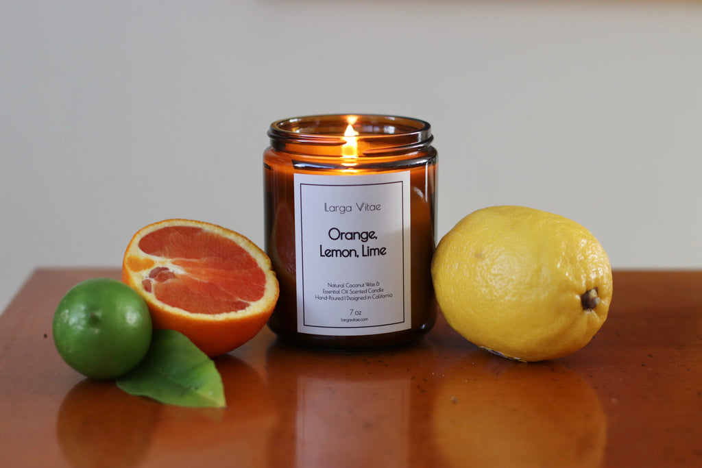 A lit Orange, Lemon, Lime Larga Vitae coconut wax essential oil candle with citrus fruits on a reflective wooden surface in front of a neutral background